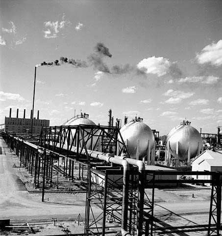Black and white photograph showing three large spherical reservoirs and a complex network of pipes in the foreground. In the background we see a tall chimney spewing out flames and smoke as well as a building with five other chimneys. 